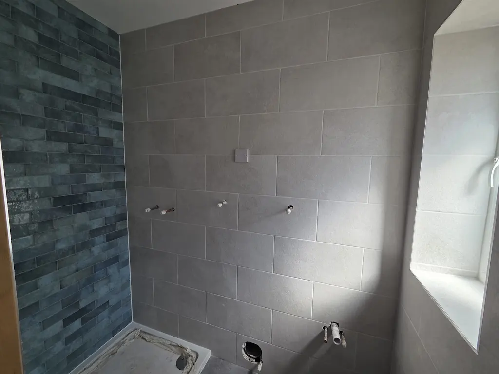 Shower recess tiled with large grey wall tiles and a dark blue brick-effect feature wall, with the tray fitted and pipework left proud.
