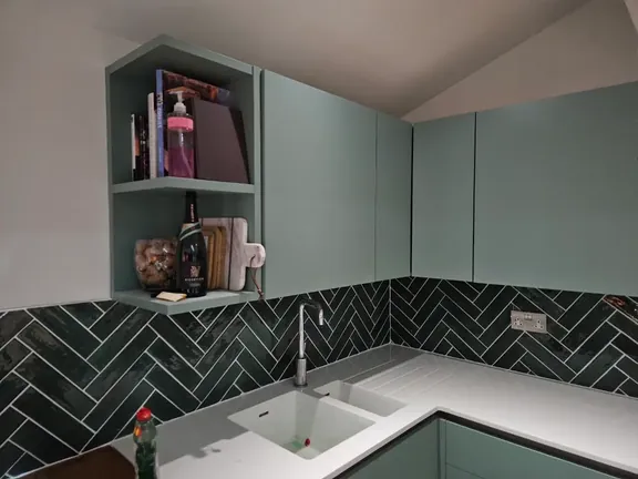 Dark green gloss tiles laid in a herringbone pattern around a corner sink run, against sage green units and a white worktop.