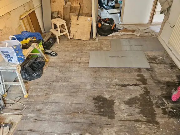 Floor stripped back to dirty, uneven original boards with building debris and tools still in the room.